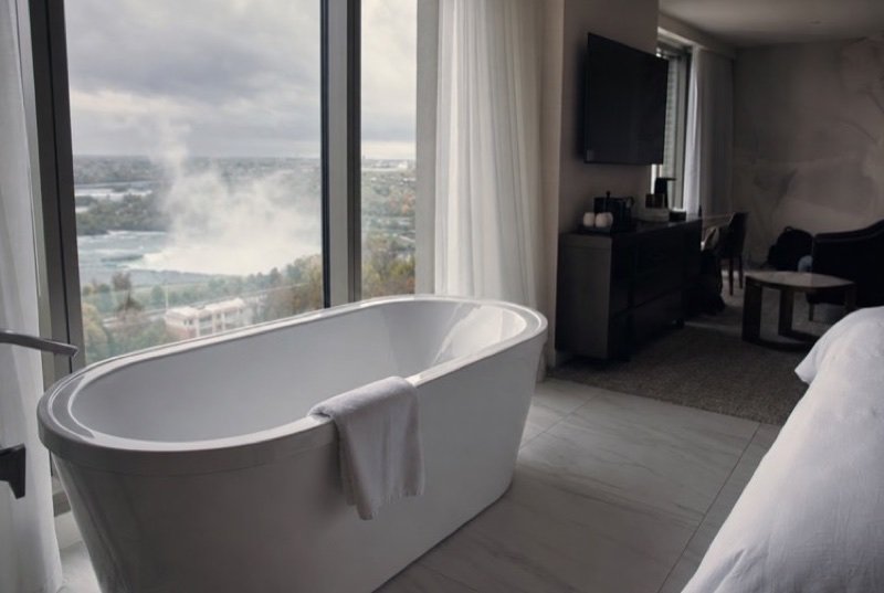 Soft daylight through sheer curtains onto a deep soaking tub set beside the window of a neat suite