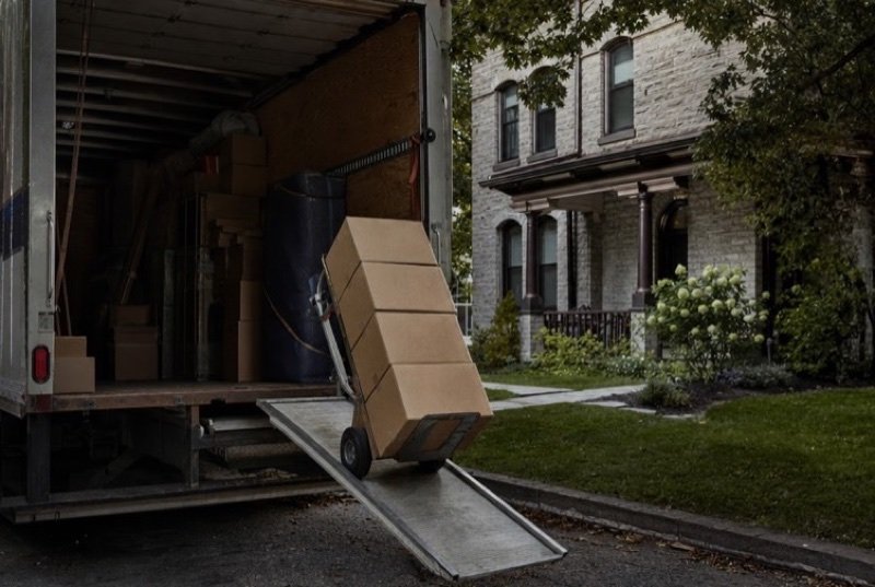 A moving truck’s ramp lowered beside a lawn, cardboard boxes piled on a hand truck, routine work in the moving industry